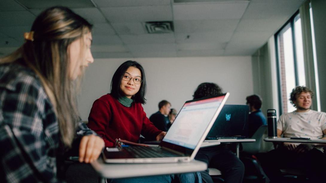 A Kettering student wearing a red sweater looks at another student. They sit in a classroom and each have open laptops